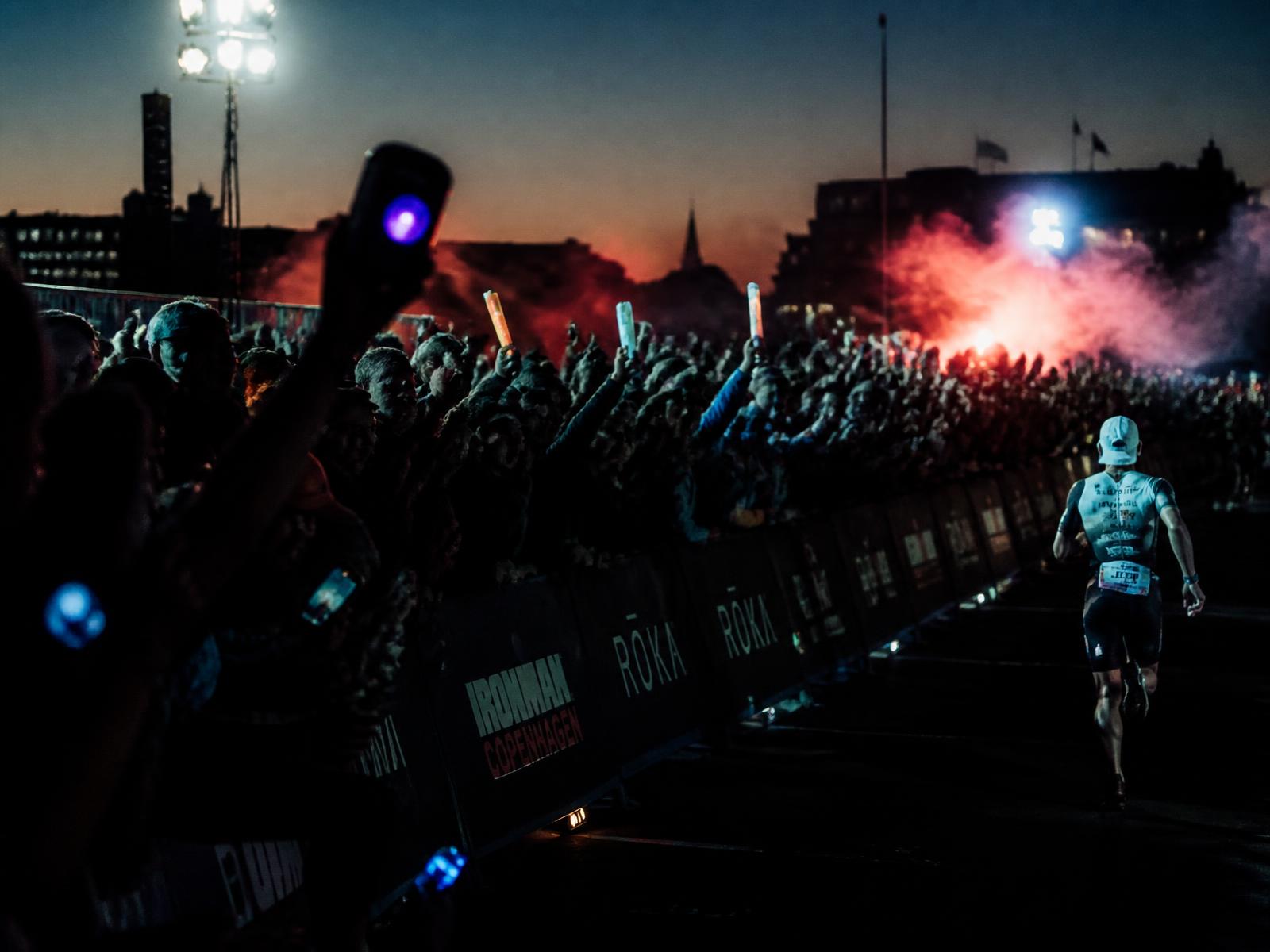 Spectators with portable speakers cheering at a night-time Ironman finish line in Copenhagen